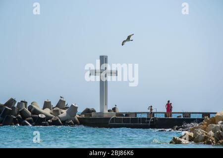 Mare, vista di una grande croce di cemento sulla baia, persone a piedi e uccelli volare, acque blu e cielo, frangiflutti, stagione estiva, natura Foto Stock