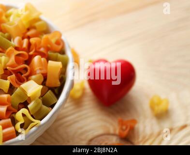 Pasta secca multicolore a forma di cuore in ciotola bianca su tavolo di legno Foto Stock