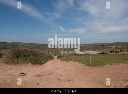 Dirt Track che conduce alla Fattoria con edifici e macchinari con un cielo blu nuvoloso luminoso sfondo in Devon Rurale, Inghilterra, Regno Unito Foto Stock