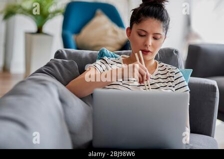 Giovane donna che mangia qualcosa con bastoncini davanti al laptop. Foto Stock