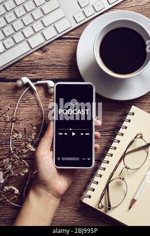concetto di contenuti audio podcast. applicazione podcast sullo schermo dello smartphone mobile su tavolo in legno con tazza da caffè, auricolari, occhiali, notebook Foto Stock