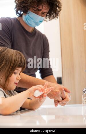 Padre caucasico indossare maschera facciale per evitare contagio insegnare bambina bambina come lavare le mani in bagno durante covid-19 pandemic blocco. Papà Foto Stock