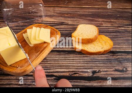 Qualcuno taglia un po' di burro con un coltello da un pezzo di burro in un piatto di burro di legno, fette di formaggio e pane tostato di grano fresco su uno sfondo di legno. C Foto Stock