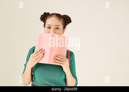 Affascinante femmina che guarda la fotocamera e nasconde metà del volto dietro il libro con copertina rosa mentre si trova su sfondo chiaro Foto Stock