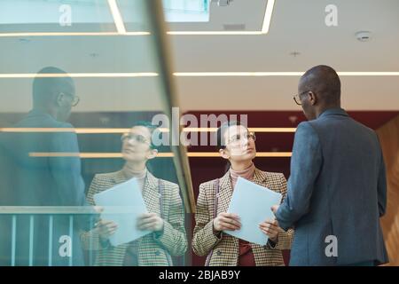Giovane imprenditore serio che tiene contratto e che parla con l'uomo d'affari africano mentre stanno in piedi al corridoio dell'ufficio Foto Stock