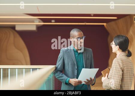 Uomo d’affari africano che parla con la giovane imprenditrice mentre si trova in ufficio corridoio che discutere di nuovo contratto Foto Stock