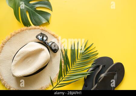 Cappello da spiaggia con paglia, infradito e foglie tropicali. Sfondo estivo con accessori su giallo. Vista dall'alto, disposizione piatta. Spazio per il testo. Foto Stock