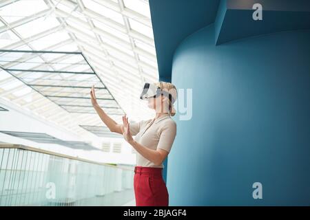 Giovane donna d'affari con capelli biondi indossando occhiali virtuali gesturing in ufficio Foto Stock
