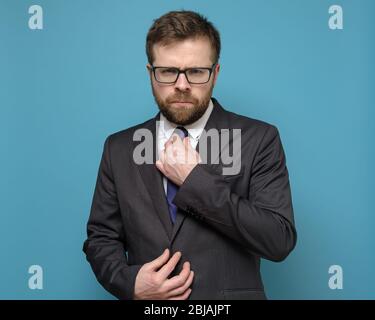 Uomo d'affari stretto e sicuro raddrizza la cravatta e con gli occhi piercing guarda nella fotocamera, su uno sfondo blu. Foto Stock