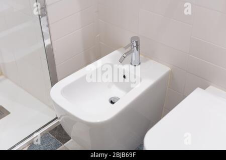 bidet bianco avvitato a una parete di piastrelle beige nel bagno, primo piano. bidet bianco con miscelatore cromato sul pavimento piastrellato Foto Stock