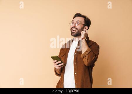 Ritratto di un giovane e allegro arttractive bearded uomo che indossa abiti casual in piedi isolato su sfondo beige, ascoltando musica con il ph mobile Foto Stock