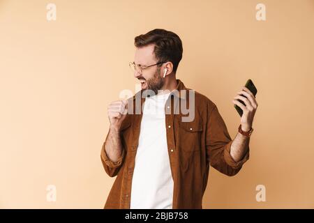 Ritratto di un giovane e allegro arttractive bearded uomo che indossa abiti casual in piedi isolato su sfondo beige, ascoltando musica con il ph mobile Foto Stock