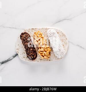 Set di tre eclair fatti in casa su sfondo di marmo. Vista dall'alto di deliziosi e sani profittoli con diversi ingredienti decorativi - cioccolato, arachidi e cocco sgranato. Quadrato Foto Stock
