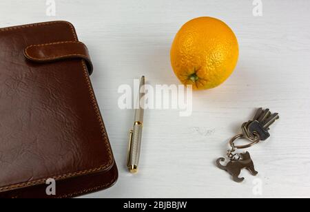 Una penna elegante, un pianificatore quotidiano con una copertina in pelle, un arancione e un set di chiavi su una scrivania in legno bianco Foto Stock