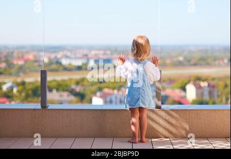 bambina carina che guarda il paesaggio della città attraverso la balaustata di vetro nel patio sul tetto Foto Stock