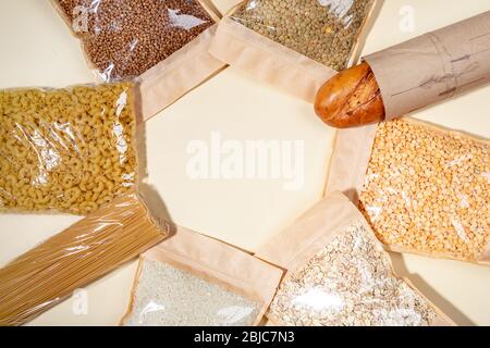 Sacchetti di carta e cellophane con cereali, legumi e pasta disposti in cerchio su beige. Grano saraceno, piselli, lenticchie, riso, spaghetti, baguette. Zero sprechi Foto Stock