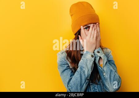 Scioccata giovane donna spaventata porga attraverso le dita, stares con gli occhi aperti ampiamente, indossa giacca e cappello denim, curioso studente femmina peek cover hi Foto Stock