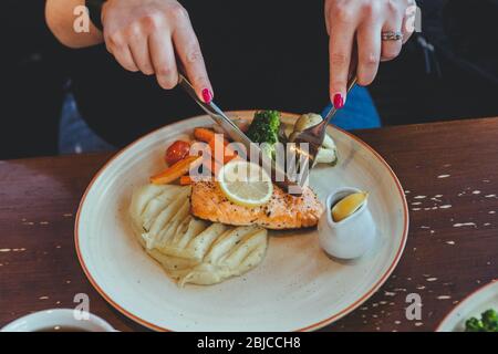 Mani di una donna seduta al tavolo in un caffè, con bistecca di pesce rosso e purè di patate con verdure su un piatto bianco per pranzo Foto Stock