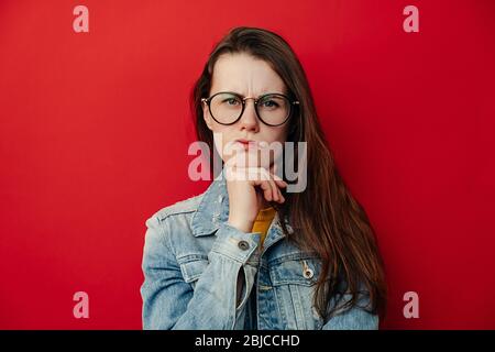 Giovane donna in occhiali curva labbra e rane sopracciglia, tiene la mano sotto il mento, guarda con espressione scontento, indossa giacca denim, si alza su rosso s. Foto Stock