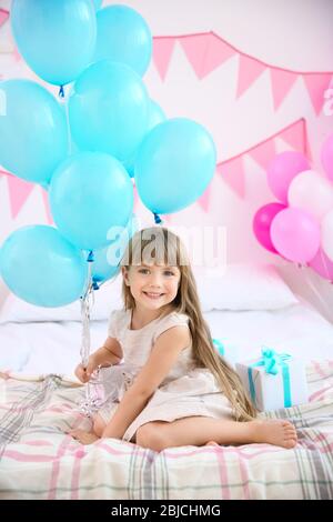 Ragazza carina con palloncini seduti a letto in camera decorata per la festa di compleanno Foto Stock