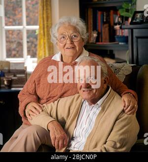 Anziani anziani anziani pensionati felice coppia 80-86 anni proprietari di casa seduti nel loro comodo soggiorno di fronte alla fotocamera Foto Stock