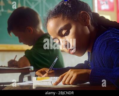 Junior ragazza scuola scolastica ragazza nera africana 9-11 anni in classe scrivendo alla sua scrivania con la geometria bionda del ragazzo e della scuola proietta dietro la lavagna Foto Stock