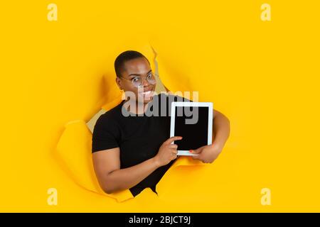 Mostra lo schermo del tablet vuoto. Allegra donna afro-americana pone in carta giallo strappato sfondo, emotivo. Rompersi, infranti. Concetto di emozioni umane, espressione facciale, vendite, annuncio. Foto Stock