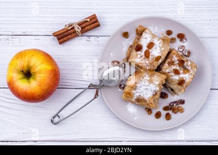 Fette di strudel di torta di mele fatta in casa in un piatto con ingredienti su un rustico tavolo di legno bianco. Torta con mele, uvetta e cannella, dolci Foto Stock