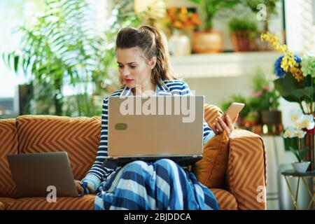 casalinga alla moda in blusa blu e giacca a righe nella casa moderna in giorno di sole utilizzando diversi dispositivi - laptop, smartphone. Foto Stock