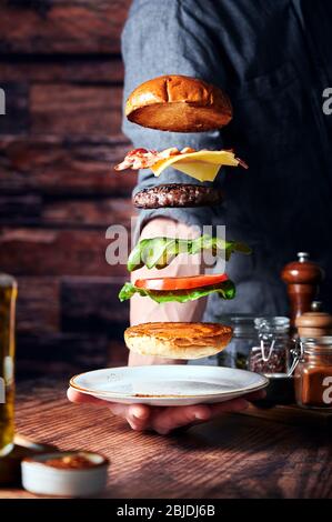 Hamburger di manzo volante con lattuga, pomodori, sottaceti aneto, polpettine di manzo, pancetta di formaggio cheddar maturo e srracha mayo. Sfondo e bancone in legno Foto Stock