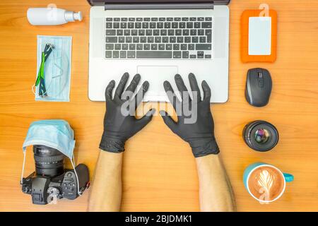 Moderno fotografo vista dal desktop durante 2020 COVID-19 su tessuto di legno. Mani maschili con guanti neri che lavorano sul laptop. Dispositivi di protezione medica Foto Stock