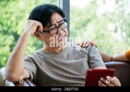 Un vecchio che sta guardando il suo tablet con il volto preoccupato o serio ottiene il sostegno dalla moglie o dalla sua famiglia Foto Stock