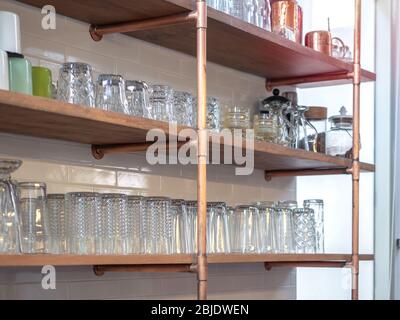 Vari bicchieri su scaffale realizzati con tubi di rame e piastre di legno su piastrelle bianche pareti in mattoni in bar. Foto Stock