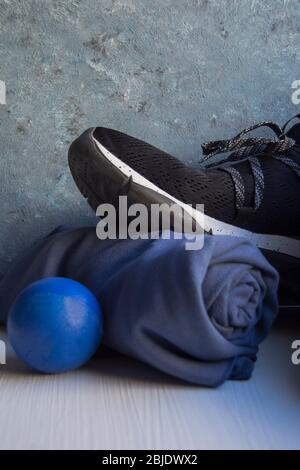 Sneaker nera con dettagli e fori bianchi, asciugamano blu arrotolato e sfera in gomma blu su base bianca e sfondo nei toni blu e grigio. Foto Stock