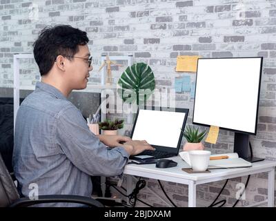 Uomo asiatico che lavora e digita con computer portatile con schermo bianco vuoto e guardando un altro monitor in camera, condominio. Lavoro a casa e busi Foto Stock