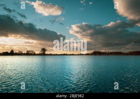 Lago turchese e tranquillo, onde dolci sull'acqua e nuvole evidenziate dal sole serale Foto Stock