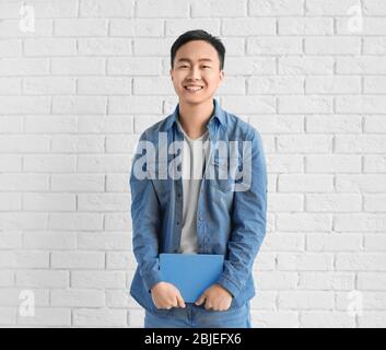 Giovane uomo asiatico con libro in piedi vicino al muro di mattoni Foto Stock