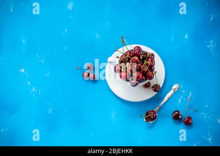 Una tazza di deliziose ciliegie dolci mature su Blue background.Composition, una tazza di ceramica bianca riempita con succose ciliegie rosse mature e diverse bacche con Foto Stock