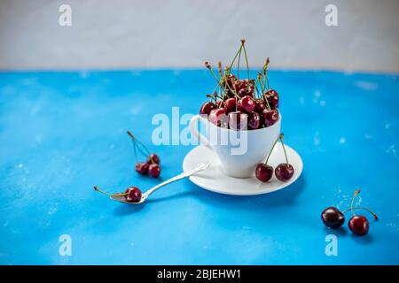 Una tazza di deliziose ciliegie dolci mature su Blue background.Composition, una tazza di ceramica bianca riempita con succose ciliegie rosse mature e diverse bacche con Foto Stock