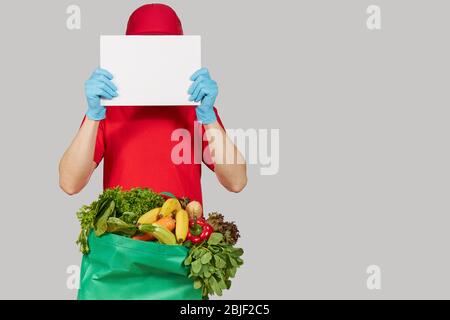 Concetto di shopping online. Corriere maschile in uniforme rossa, maschera protettiva e guanti con una scatola di alimentari con frutta fresca e verdure tiene un divieto bianco Foto Stock