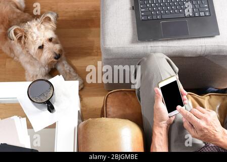 Primo piano delle mani di un uomo con il telefono cellulare che lavora a casa durante il blocco Covid-19. Colpo ad angolo alto con cane da compagnia ai suoi piedi. Foto Stock