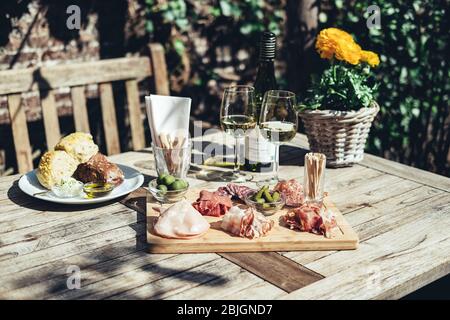 Salumi serviti con vino bianco all'aperto. Un piatto di prosciutto e pancetta affettare con sottaceti su una tavola di legno Foto Stock