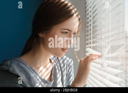 Bella ragazza giovane che separa le stecche di ciechi e guardando attraverso la finestra Foto Stock