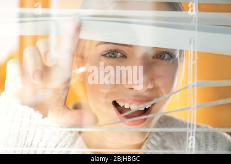 Bella ragazza giovane che separa le stecche di ciechi e guardando attraverso la finestra Foto Stock