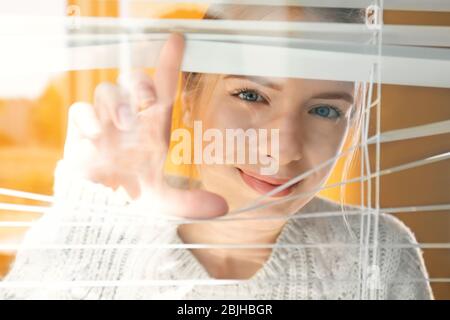 Bella ragazza giovane che separa le stecche di ciechi e guardando attraverso la finestra Foto Stock