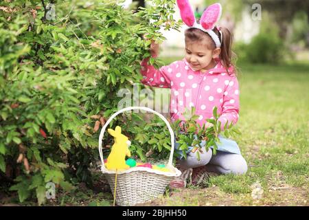 Ragazza carina con cesto vicino Bush nel parco. Concetto di caccia all'uovo di Pasqua Foto Stock