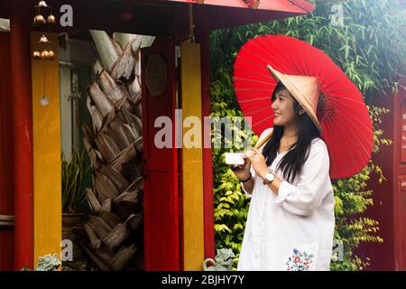 Viaggiatori donne thailandesi che indossano abiti etnici vietnamiti con cappello di bambù e ombrello rosso per scattare foto al giardino e vintage retro casa in reso Foto Stock