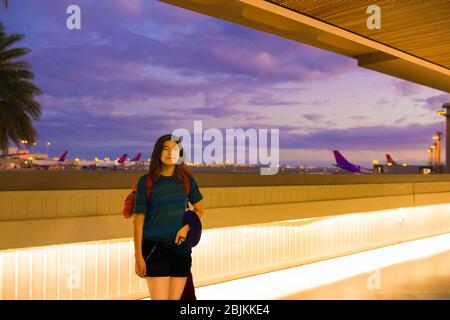 Adolescente con zaino rosso in piedi lungo il corridoio esterno all'aeroporto Hawaiiano prima di prendere il volo serale. Aerei e cieli colorati a bac Foto Stock