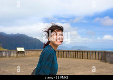 Giovane ragazza caucasica asiatica biraciale o giovane adulto che sta al Pali Lookout, Oahu, Hawaii in una giornata soleggiata e ventosa con vista della città di Kaneohe e dell'oceano blu Foto Stock