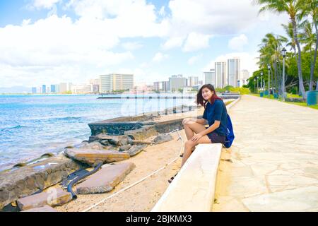 Sorridente giovane donna o ragazza biraciale asiatica Cuacasiana seduta lungo il sentiero di pietra lungo le acque di Waikiki a Honolulu, Hawaii, Oahu. Hote Foto Stock